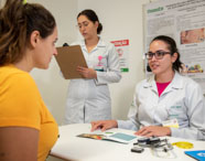 Atendimento nutricional na clínica-escola: estudante de Nutrição, de jaleco branco, orienta paciente sentada à mesa, mostrando folheto educativo. Outra aluna faz anotações em prancheta. Ambiente limpo, com cartaz informativo ao fundo e instrumentos como fita métrica e adipômetro sobre a mesa.