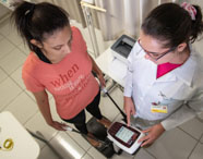 Vista superior de avaliação de bioimpedância: paciente em camiseta coral segura eletrodos conectados a aparelho moderno, acompanhada por estudante de Nutrição em jaleco branco. No chão, balança, fita métrica e outros equipamentos típicos de consultório nutricional.