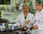 Duas profissionais ou estudantes de Nutrição preparam carne em cozinha experimental. Ambas usam jaleco branco e touca estampada, uma delas mexe cubos de carne em frigideira no fogão industrial, com panelas de inox ao lado. Ambiente amplo, típico de laboratórios de gastronomia universitária