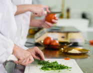 Close de mãos de estudantes de Nutrição cortando cheiro-verde em tábua branca, com tomates e balança ao fundo. Todos usam jaleco branco, evidenciando atividade prática de preparo de alimentos em laboratório culinário universitário.