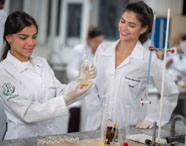 Estudantes de Nutrição em laboratório de bioquímica, usando jaleco branco e luvas, analisam líquidos em frascos de vidro. Uma aluna segura erlenmeyer com solução amarela, outra observa. Bancada de granito com tubos de ensaio e bureta, ambiente acadêmico e científico.