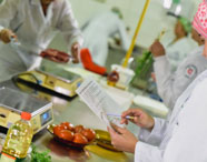 Alunos de Nutrição em laboratório de análise de alimentos, um deles consulta ficha técnica enquanto outros manipulam ingredientes como carne, tomates e ervas frescas. Balanças digitais, recipientes e utensílios espalhados sobre bancada de inox, ilustrando rotina prática do curso.