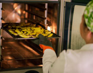 Estudante de Nutrição com touca verde coloca bandeja cheia de batatas temperadas em forno industrial. O ambiente é de cozinha experimental, com iluminação quente e utensílios profissionais, representando a prática de preparo saudável de alimentos no curso universitário.