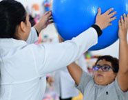 Aluna do curso de Terapia Ocupacional auxiliando paciente infantil em exercício de reabilitação com bola azul, demonstrando prática clínica e cuidado.