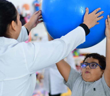 Aluna do curso de Terapia Ocupacional auxiliando paciente infantil em exercício de reabilitação com bola azul, demonstrando prática clínica e cuidado.