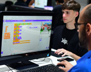 Aluno do curso de Sistemas de Informação programa em ambiente visual, com blocos coloridos no monitor Dell. O código representa lógica de programação e há uma animação de personagem ao lado de uma casa. Ao fundo, outro estudante acompanha a atividade, ilustrando o ensino prático em laboratório da instituição.