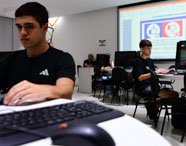 Estudantes de Sistemas de Informação participam de aula prática em laboratório moderno, cada um utilizando computadores Dell. No fundo, projetor exibe slide com ícones de positivo e negativo. Ambiente claro, mesas distribuídas em grupos, reforçando o trabalho colaborativo típico do curso na instituição.