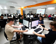 Visão ampla do laboratório de Sistemas de Informação, com diversos estudantes distribuídos em mesas redondas, todos utilizando computadores Dell. O ambiente é iluminado, com detalhes em laranja e decoração geométrica nas paredes, promovendo integração e aprendizado coletivo em tecnologia.