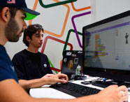 Dois alunos do curso de Sistemas de Informação compartilham mesa, programando em ambiente visual de blocos. Um utiliza notebook personalizado com adesivos, enquanto o outro digita no teclado do desktop. A parede traz desenhos coloridos, reforçando o ambiente criativo da instituição.