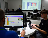 Aluno do curso de Sistemas de Informação trabalha em dupla, programando em monitor Dell com interface de blocos. Outro estudante acompanha a atividade. Ao fundo, projeção de conteúdo didático na parede, destacando a metodologia ativa e colaborativa do curso em laboratório universitário.