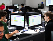 Laboratório de criação em Sistemas de Informação, com vários estudantes concentrados em seus computadores. Monitores exibem códigos em blocos, simulando projetos de lógica e jogos. Ambiente amplo, mesas brancas e decoração moderna, ideal para o desenvolvimento de habilidades técnicas na faculdade.