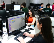 Alunas do curso de Sistemas de Informação sentadas em mesa redonda, programando em computadores Dell. Na tela, interface de blocos coloridos e animação de personagem. Celulares sobre a mesa, ambiente descontraído e colaborativo, característico das aulas práticas da instituição.