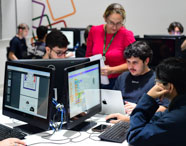 Professora do curso de Sistemas de Informação acompanha grupo de alunos em laboratório, auxiliando-os em tarefas de programação. Alunos utilizam computadores Dell e notebooks Apple, enquanto a docente observa e esclarece dúvidas. Ambiente dinâmico, com decoração colorida e atmosfera colaborativa.