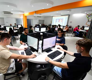 Visão geral do laboratório de Sistemas de Informação em Prudente, com estudantes distribuídos em mesas redondas, cada um em frente a computadores Dell. O espaço é amplo, bem iluminado, com detalhes em laranja e decoração geométrica, representando o ambiente moderno e tecnológico da instituição.