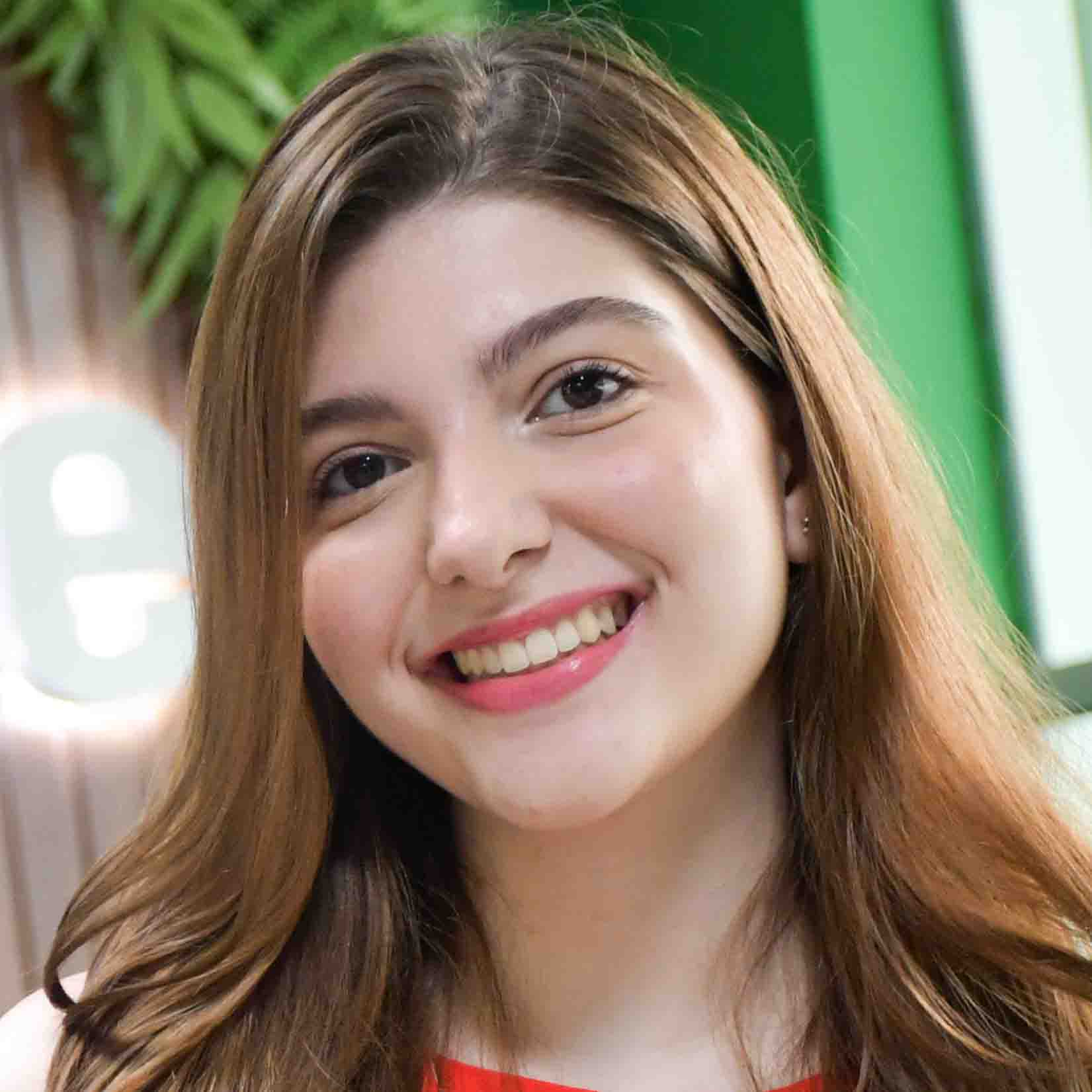 Giovanna, aluna do curso de fonoaudiologia, trajando uma blusa branca com detalhes em vermelho. Ela está em um ambiente interno com fundo composto por elementos verdes e madeira, sugerindo um espaço institucional ou decorativo. A iluminação suave destaca os cabelos castanhos claros e o contraste com o cenário, criando uma composição acolhedora e harmoniosa.