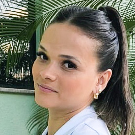 Julyssandra, aluna do curso de fonoaudiologia, trajando um jaleco branco. Ela está em um ambiente com fundo formado por folhagens verdes e uma parede clara, sugerindo um espaço acadêmico. A iluminação natural realça os detalhes do cabelo preso em um rabo de cavalo, transmitindo um ar profissional e sereno.