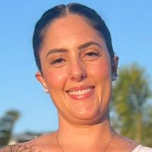 Mayara, aluna do curso de fonoaudiologia, trajando uma blusa branca. Ela está em um ambiente externo com fundo azul do céu e árvores ao longe, sugerindo um dia ensolarado em um espaço aberto. A iluminação natural destaca os detalhes do colar dourado e cria um contraste vibrante com o cenário, transmitindo leveza e descontração.