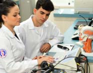 Dois estudantes de Fonoaudiologia, em jalecos brancos, operam equipamento de audiometria em laboratório. Sobre a bancada, há modelo anatômico de cabeça e microfone, ilustrando a integração entre teoria e prática no processo de avaliação auditiva.