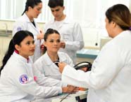 Grupo de estudantes de Fonoaudiologia, todos de jaleco branco, recebem orientações de professora durante aula prática em laboratório. O ambiente conta com equipamentos e materiais didáticos, reforçando o caráter formativo e interativo da instituição.