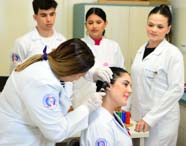 Professora de Fonoaudiologia examina ouvido de estudante com otoscópio, usando luvas e jaleco branco. Outros alunos acompanham o procedimento, atentos, em sala equipada com materiais didáticos, reforçando o aprendizado prático e supervisionado do curso.