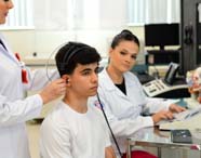 Estudante de Fonoaudiologia posiciona fones de ouvido e plug auricular em paciente para teste auditivo, enquanto colega opera aparelho de audiometria. Laboratório conta com modelos anatômicos de cabeça e equipamentos modernos, promovendo prática realista na Unoeste.