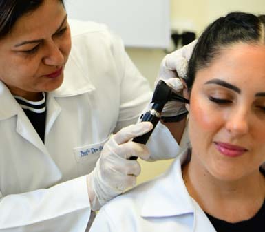 Em ambiente clínico, profissional ou estudante do curso de Fonoaudiologia utiliza otoscópio para examinar o ouvido de um paciente. Ambos vestem jaleco branco e luvas descartáveis, evidenciando prática segura e detalhada na avaliação auditiva, típica de atendimentos acadêmicos em instituição de ensino.