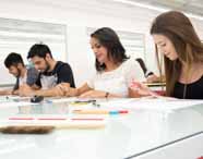 Grupo de quatro estudantes do curso de Design de Interiores sentados lado a lado em sala iluminada, praticando desenho em folhas brancas sobre mesa de vidro. Diversos materiais como canetas coloridas, borracha e pincel estão espalhados à frente. Ambiente moderno e colaborativo de instituição de ensino