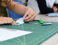 Mãos femininas com unhas pintadas de verde água dobram papel branco sobre base de corte quadriculada, ao lado de estilete verde e régua metálica. Cena típica de atividade prática manual do curso de Design de Interiores, evidenciando atenção aos detalhes e habilidades artesanais dos estudantes.