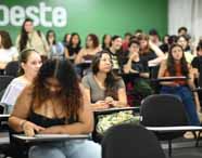 Turma numerosa de estudantes sentados em carteiras individuais, atentos a aula teórica em sala ampla com parede verde e letreiro parcial da instituição Unioeste ao fundo. Diversidade de roupas e estilos entre os alunos, representando ambiente acadêmico inclusivo do curso de Design de Interiores.