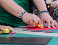 Duas pessoas cortam papel vermelho com estiletes amarelos sobre base de corte verde. Ambas usam pulseiras coloridas e camisa verde. Cena evidencia atividade prática manual, comum nas aulas do curso de Design de Interiores, onde precisão e criatividade são essenciais para os estudantes.