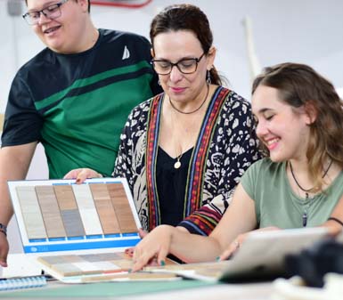 Grupo de estudantes e professora do curso de Design de Interiores analisam amostras de revestimentos em catálogo aberto sobre a mesa. O ambiente é de sala de aula, com materiais de apoio, clima colaborativo e foco na escolha de texturas e cores para projetos de interiores na Unoeste.