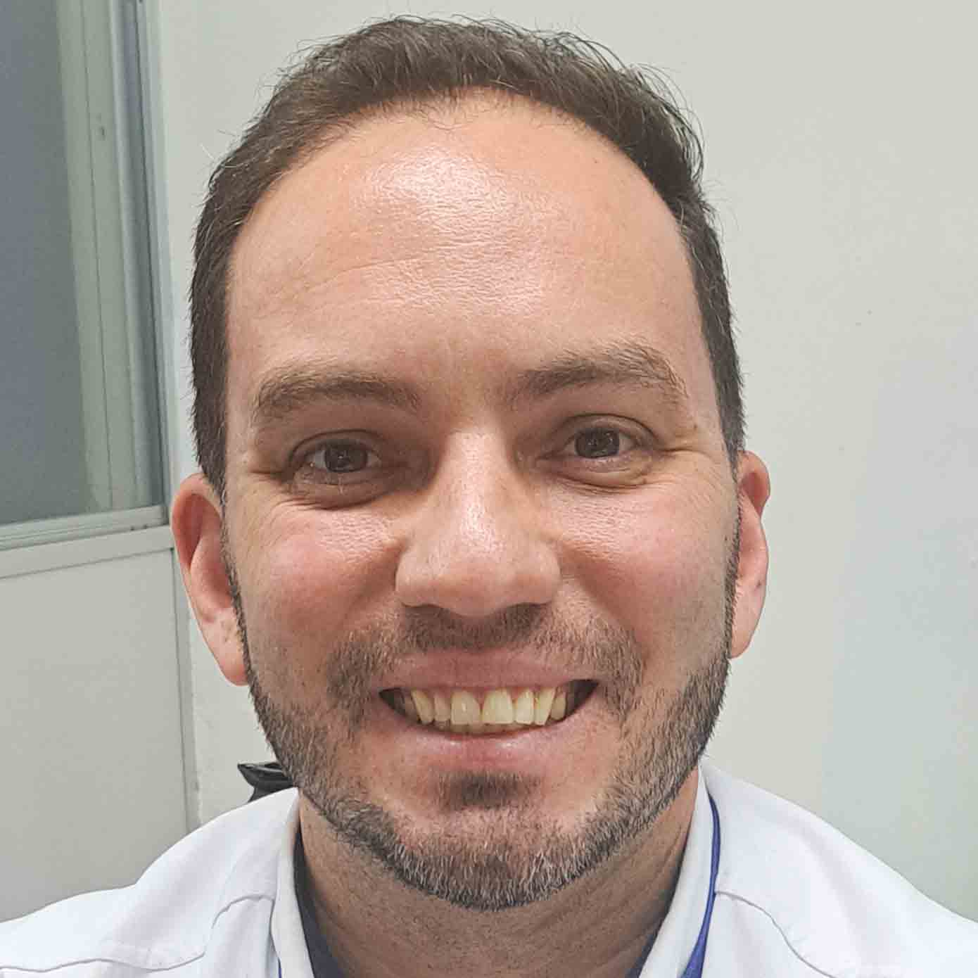 Carlos, aluno do curso de gestão comercial, em um ambiente interno com uma parede clara e janela ao fundo. Ele veste uma camisa branca que complementa o cenário neutro.
