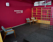Sala de convivência da Business School Unoeste, com paredes vermelhas, prateleiras de madeira, cadeiras estofadas cinza claro e divisória metálica. Espaço acolhedor e moderno para encontros e estudos dos alunos de Gestão Comercial.
