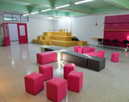 Ambiente amplo e moderno com pufes quadrados rosa e cinza, arquibancada de madeira clara, cadeiras pretas alinhadas e luminárias pendentes. Sala de convivência e integração para atividades colaborativas do curso de Gestão Comercial na Unoeste.