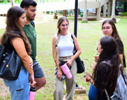 Grupo de cinco estudantes conversa em círculo sobre gramado no campus da Unoeste, em Presidente Prudente. Eles usam mochilas, garrafas reutilizáveis e roupas casuais, demonstrando integração e vida universitária no curso de Gestão Comercial.
