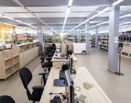 Biblioteca ampla e moderna da Unoeste, com mesas de atendimento equipadas com computadores, cadeiras giratórias e estantes repletas de livros organizados. Ambiente iluminado, ideal para pesquisa e estudo dos alunos de Gestão Comercial.
