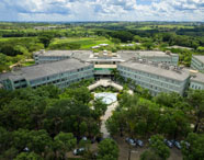 Vista aérea do campus Unoeste em Presidente Prudente, mostrando edifícios dispostos em formato semicircular cercados por áreas verdes, árvores e jardins. Ao centro, fonte circular e bandeiras, evidenciando a grandiosidade da instituição.