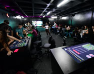 Laboratório inovador da Unoeste com mesas digitais interativas, luzes coloridas e estudantes utilizando recursos tecnológicos. Ambiente escuro, propício para simulações e atividades práticas do curso de Gestão Comercial, promovendo aprendizado dinâmico.