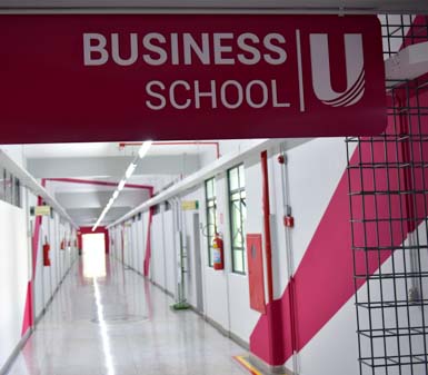 Corredor interno moderno da Business School Unoeste, com destaque para uma placa suspensa vermelha e branca indicando "Business School U". As paredes brancas possuem detalhes geométricos em vermelho, janelas de vidro e extintores, compondo ambiente acadêmico do curso de Gestão Comercial em Presidente Prudente.
