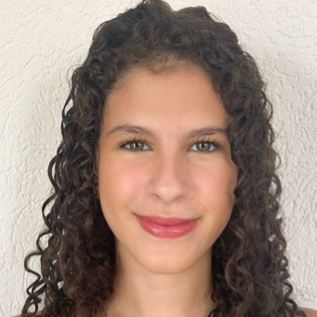 Maria Beatriz, aluna do curso de arquitetura e urbanismo, em um ambiente interno com uma parede texturizada ao fundo. Seu cabelo cacheado escuro complementa a cena.