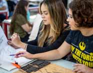 Faculdade de Arquitetura em Presidente Prudente: duas alunas analisam e discutem projetos arquitetônicos impressos, utilizando régua e materiais sobre a mesa. O ambiente de sala de aula evidencia a prática e o desenvolvimento de competências técnicas do curso.