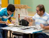 Faculdade de Arquitetura em Presidente Prudente: aluno e professor realizam atividade prática de montagem de estruturas, utilizando maquetes físicas e instrumentos de medição. O espaço de laboratório destaca a vivência prática essencial à formação do arquiteto.