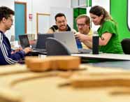 Faculdade de Arquitetura: quatro pessoas, sendo três homens e uma mulher, sentam-se ao redor de uma mesa branca usando notebooks prateados da Dell, analisando projetos. Ao fundo, uma parede verde vibrante destaca o ambiente moderno. No primeiro plano, há peças de madeira desfocadas, sugerindo modelos de maquete.