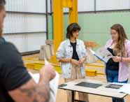 Faculdade de Arquitetura: dois alunos, um de camisa listrada azul e outro de óculos, trabalham juntos em uma mesa, montando estruturas metálicas em miniatura. O ambiente tem fundo industrial, com vigas amarelas, concreto e outros alunos ao fundo, indicando prática de engenharia estrutural.