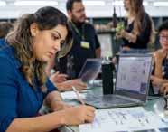 Faculdade de Arquitetura: jovem mulher de cabelo castanho e blusa azul escura escreve em caderno ao lado de notebook aberto com projetos na tela. Ao fundo, outros alunos trocam ideias em um laboratório com luz branca intensa, peças de maquete e plantas de arquitetura espalhadas pelas mesas.