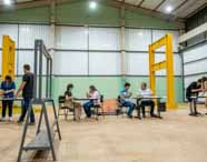 Faculdade de Arquitetura: imagem de laboratório amplo com piso claro e paredes verdes, destacando estruturas metálicas amarelas de grande porte. Grupos de alunos, trabalham em mesas montando modelos estruturais. O ambiente é organizado, iluminado e voltado à prática acadêmica.