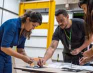 Faculdade de Arquitetura: alunos e professor interagem juntos em uma mesa, montando peças metálicas para modelos estruturais. Uma mulher de macacão azul escuro trabalha atenta ao lado de um homem de camiseta preta com crachá verde. Ao fundo, estrutura amarela reforça o ambiente de laboratório prático.