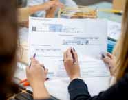 Faculdade de Arquitetura: mãos femininas seguram folha A3 com projeto arquitetônico detalhado em tons de azul e cinza, mostrando elevação de fachadas. Canetas e lápis coloridos, além de uma maquete de palitos de madeira desfocada ao fundo, reforçam o clima de estudo e criação em arquitetura.