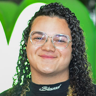 Adrian, aluno do curso de administração, aparece com cabelo cacheado e longo, em tons escuros com mechas claras nas pontas. Ele veste uma camiseta preta com uma escrita em branco na gola. O fundo é composto por um painel verde vibrante com detalhes brancos curvados, que criam contraste com sua vestimenta e destacam o cenário ao redor. A iluminação é clara, evidenciando os elementos visuais da imagem. 