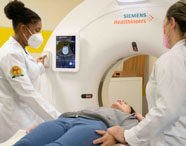 Estudantes do curso de Radiologia auxiliam paciente durante exame de tomografia computadorizada em laboratório da instituição. O paciente está deitado na mesa do equipamento Siemens Healthineers, enquanto as alunas, de jaleco branco, monitoram o procedimento e observam a tela com imagem médica detalhada.